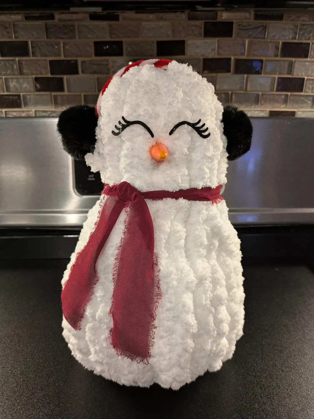 A snow lady created from finger-knitted fabric. It stands at about 1ft tall. It has a red scarf, eyelashes, candy-cane earmuffs, and a 'carrot' nose.