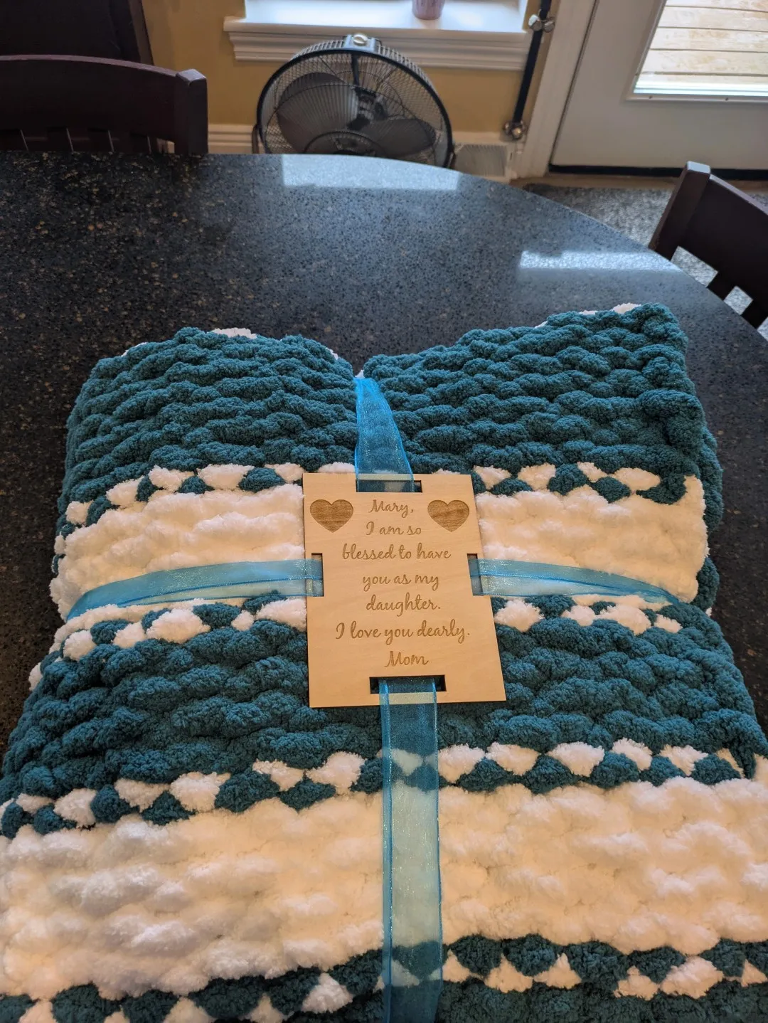A blanket made from white, and dark turquoise fabric. It is folded and wrapped, held together by a wooden plaque with text.