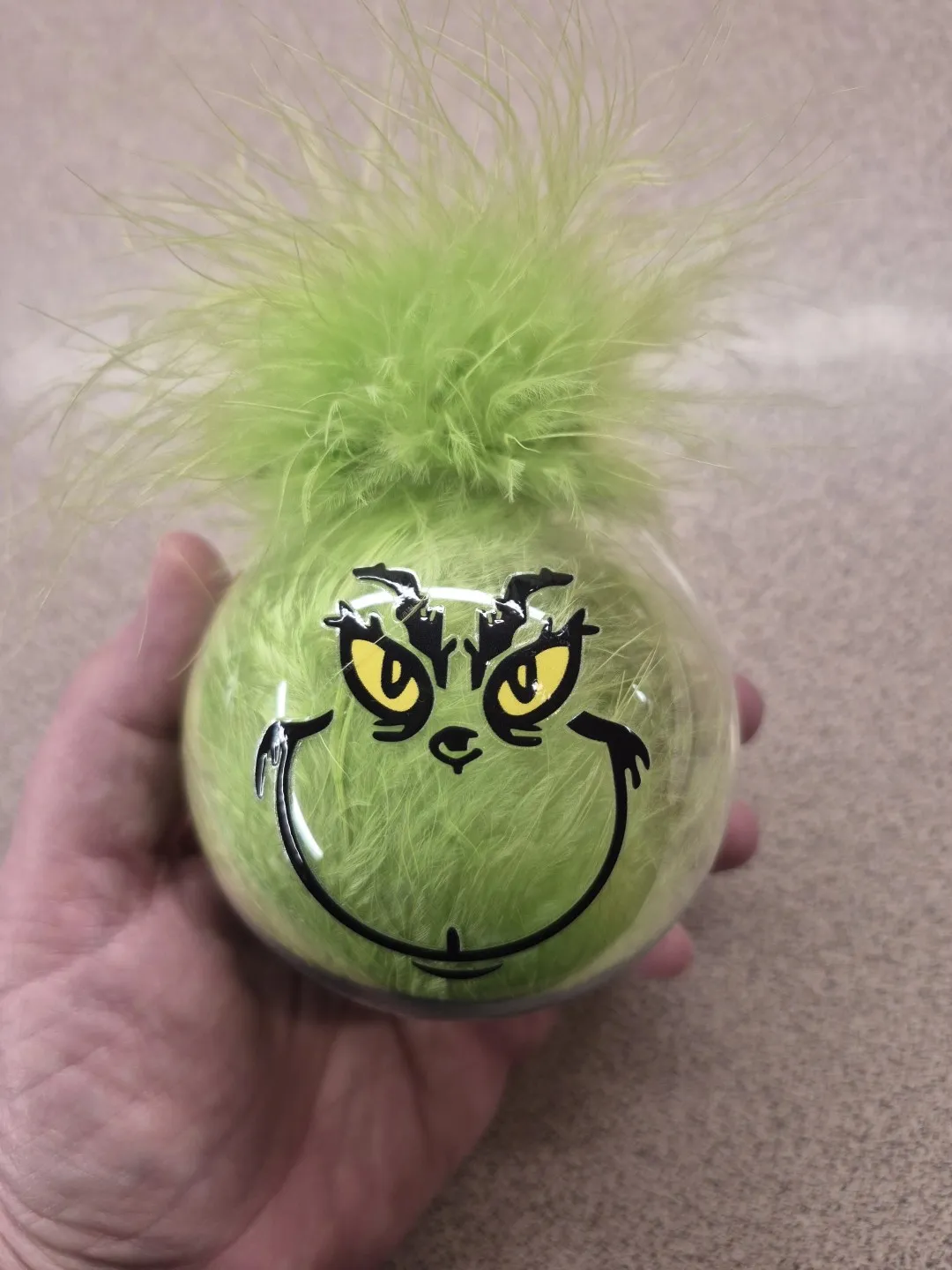 A spherical ornament with the Grinch's face on it. Inside, is green fur, with some poking out above the top.