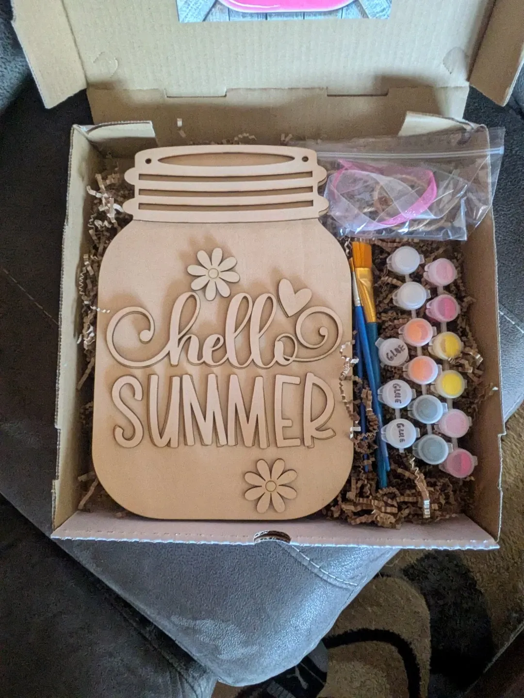 A DIY kit in a box. There is a wood piece engraved to look like a jar, which has the words 'Hello Summer' on it with some flowers. Next to it are some paint, paintbrushes, rubber bands, and packing materials.