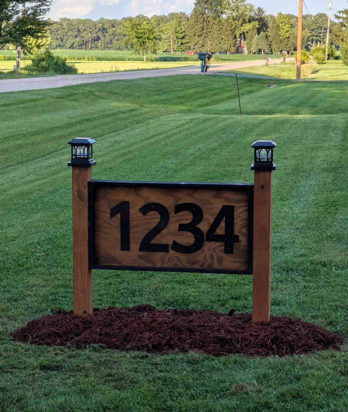 A rustic wooden home address sign displaying the black numbers 1234 between two wooden posts topped with black lanterns, photographed in a green front yard.