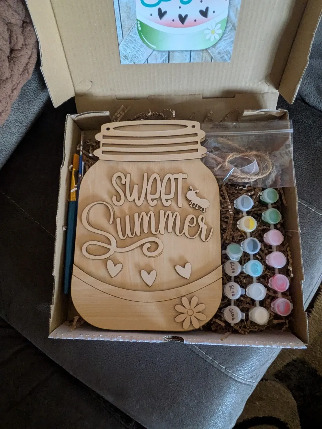 A DIY kit in a box with a wood piece engraved to look like a jar reading 'Sweet Summer' with flowers, plus paint, brushes, and packing materials.